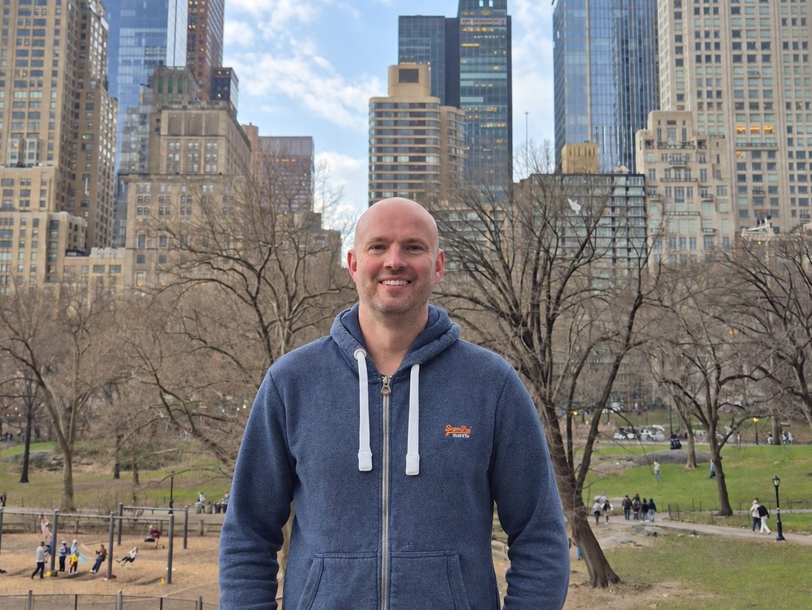 Chris smiling in Central Park with New York City buildings behind him.